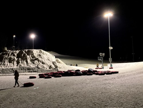 A snowy nighttime scene with red snow tubes scattered on the ground and illuminated by bright lights.