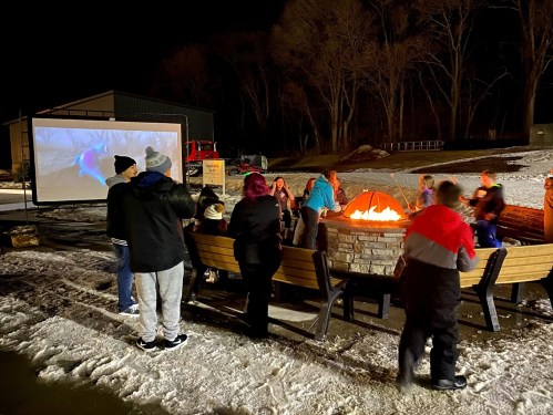 A group of people gathers around a fire pit at night, with a large screen showing a movie in the background.