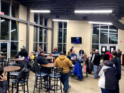 A crowded indoor space with people gathered around tables, socializing and enjoying refreshments. Large windows and a TV visible.