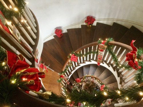 A spiral staircase decorated with garlands, red bows, and poinsettias, illuminated by soft lights.