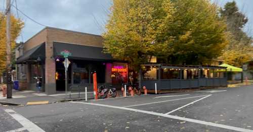 A street view of a restaurant with outdoor seating, surrounded by autumn trees and a cloudy sky.