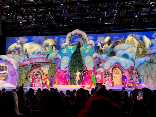Colorful stage set with whimsical buildings and a large Christmas tree, featuring performers in festive costumes. Audience in foreground.