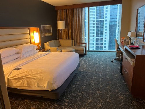 A modern hotel room featuring a king-sized bed, armchair, desk, and large window with city views.