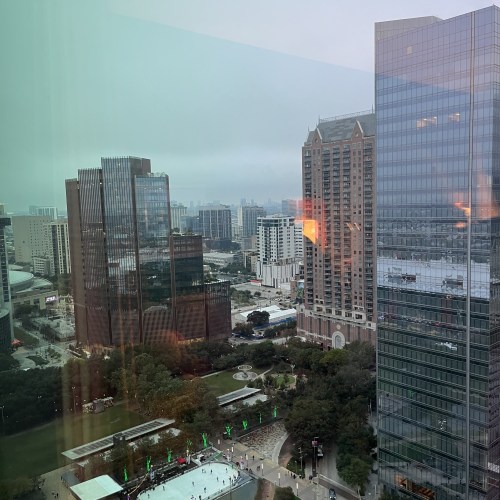 View of a city skyline with modern buildings and a park, seen through a glass window on a cloudy day.