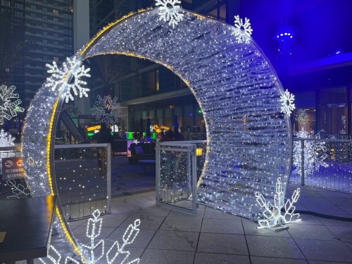 A festive archway adorned with white lights and snowflake decorations, set in a vibrant, illuminated outdoor space.