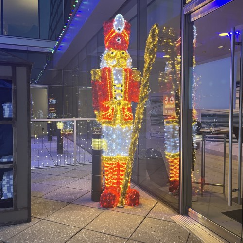 A brightly lit nutcracker figure stands outside a building, decorated with colorful lights against a twilight backdrop.
