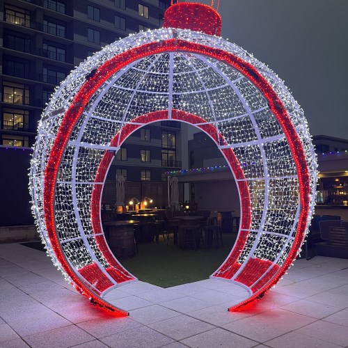 A large, illuminated ornament structure with red and white lights, set in an outdoor area with tables and chairs.