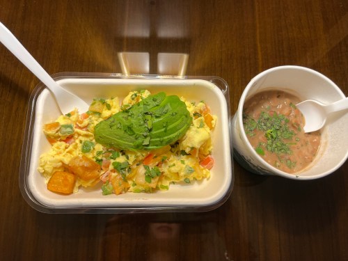 A takeout container with scrambled eggs, avocado, and vegetables, alongside a cup of refried beans topped with herbs.