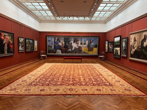 A spacious art gallery featuring framed paintings on red walls and a large ornate rug in the center.