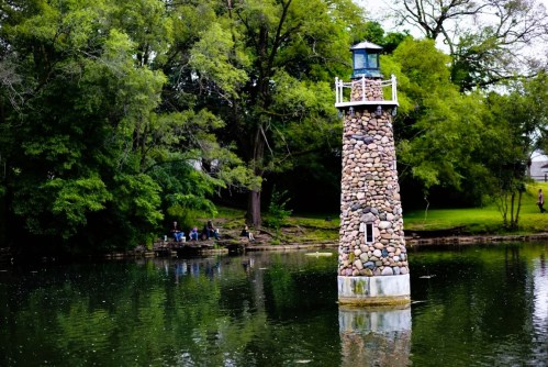 A stone lighthouse stands by a calm lake, surrounded by lush greenery and people sitting on the shore.
