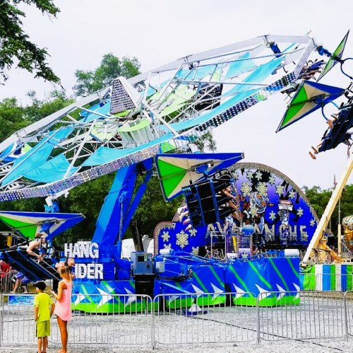 A colorful amusement ride called "Hang Glider" at a fair, with children watching and trees in the background.