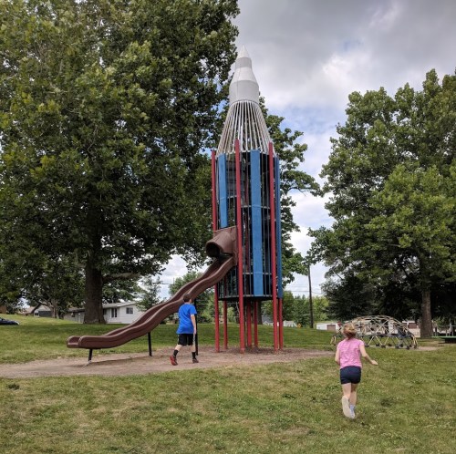 Children play near a rocket-shaped slide in a park, surrounded by trees and grassy areas.