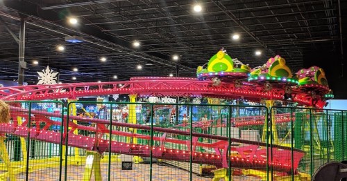A colorful indoor roller coaster with bright lights and a fenced area, surrounded by arcade games.