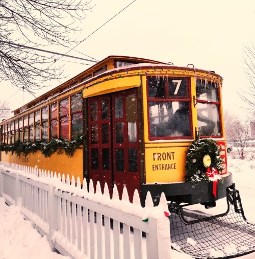A vintage yellow streetcar decorated for the holidays, surrounded by snow and a white picket fence.