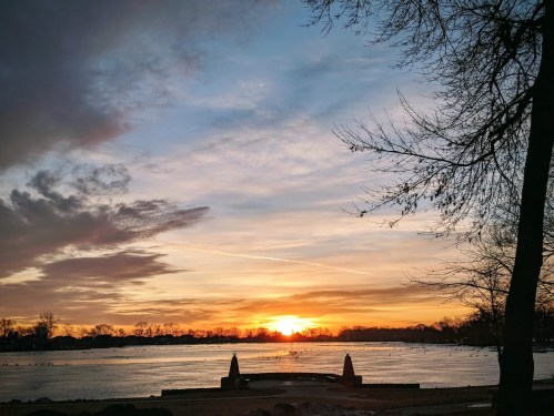 Sunset over a frozen lake, with silhouettes of trees and a stone structure in the foreground against a colorful sky.