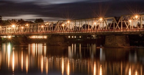 A lit bridge spans a calm river at night, with reflections shimmering on the water's surface.