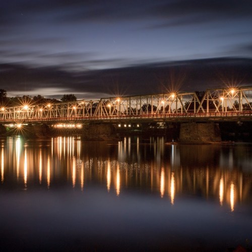 A lit bridge spans a calm river at dusk, with reflections shimmering on the water's surface.