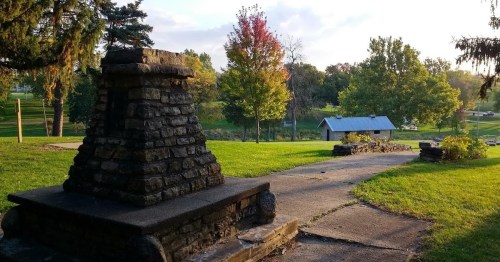 A stone structure in a park with a pathway, surrounded by trees and a glimpse of a building in the background.