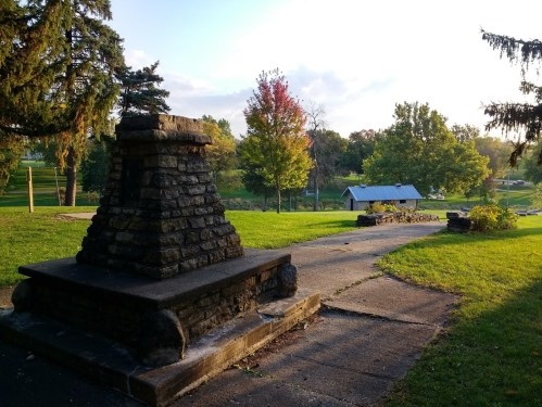 A stone structure beside a pathway in a park, with trees and a shelter in the background under a clear sky.