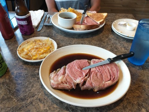 A plate with a large slice of prime rib in gravy, accompanied by a side of hash browns and a drink in a blue glass.