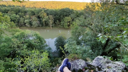 A person's feet in sneakers resting on a rock, overlooking a serene river surrounded by lush greenery.