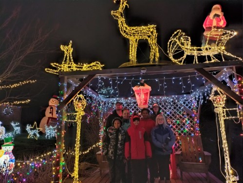 A group of people stands under a decorated gazebo with festive lights and holiday decorations, including Santa and reindeer.