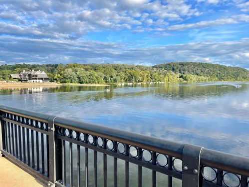 A serene river scene with lush green hills and a cloudy blue sky, viewed from a black railing.