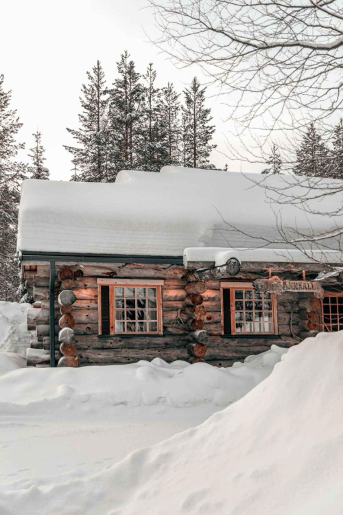 A cozy log cabin covered in snow, surrounded by tall trees in a winter landscape.