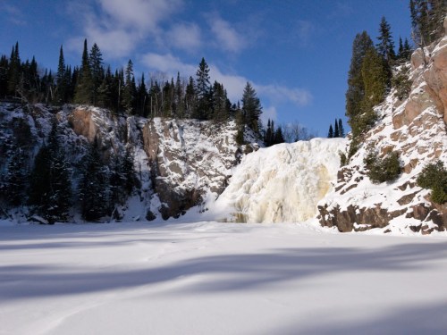 Frozen waterfall cascading down rocky cliffs, surrounded by snow-covered ground and evergreen trees under a blue sky.