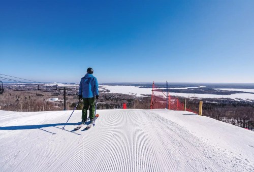 A skier stands at the top of a snowy slope, overlooking a vast landscape under a clear blue sky.