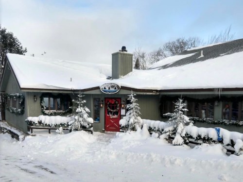 A cozy, snow-covered cabin with a red door, surrounded by evergreen trees and winter decorations.