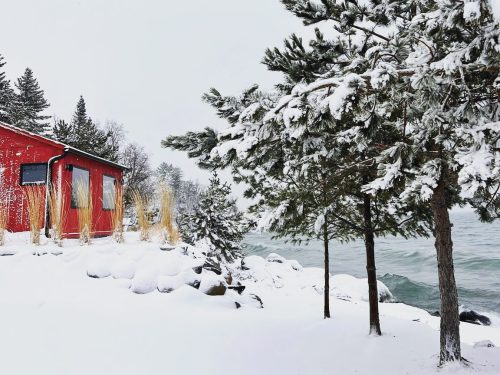 A red cabin by a snowy shoreline, surrounded by evergreen trees and a calm, icy lake.