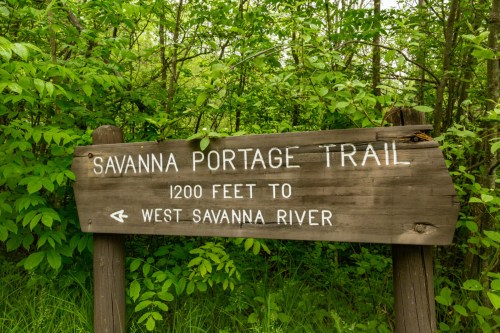 Wooden sign for Savanna Portage Trail, indicating 1200 feet to West Savanna River, surrounded by greenery.