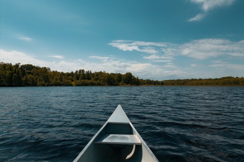 A canoe's bow on a calm lake, surrounded by lush green trees under a clear blue sky.