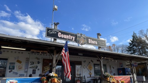 A rustic country store with an American flag, surrounded by autumn decorations and a clear blue sky.