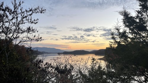 A serene sunset over a lake, framed by trees and mountains, with soft clouds in the sky reflecting on the water.