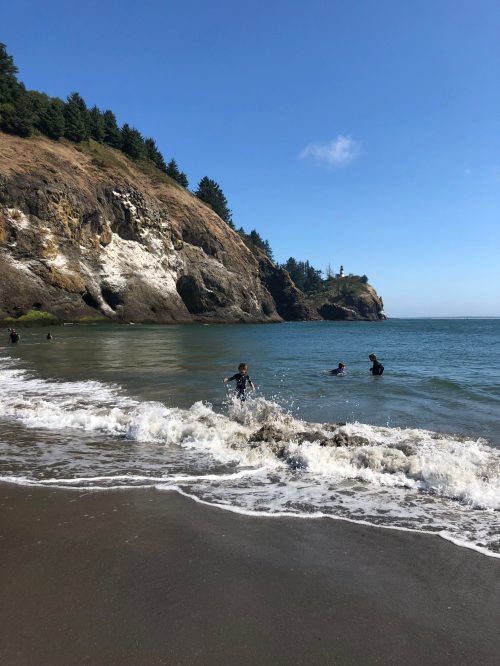 A beach scene with people playing in the water, rocky cliffs, and trees in the background under a clear blue sky.