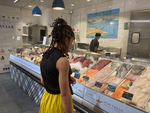 A person with braided hair looks at a seafood display in a market, featuring various fish and seafood options.