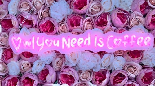 A pink sign that reads "All You Need Is Coffee" against a backdrop of pink and white roses.
