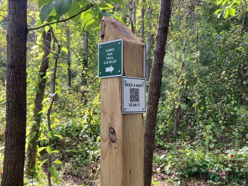 Trail sign for Hawks Run Trail, indicating a distance of 0.12 miles, with a QR code for a map. Surrounded by greenery.
