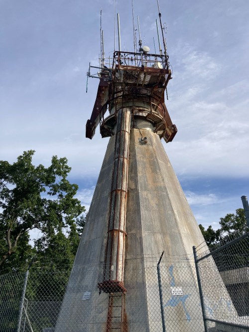 A tall, weathered communication tower with antennas, surrounded by a chain-link fence and trees in the background.
