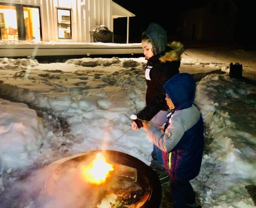 Two children roast marshmallows over a fire pit in a snowy outdoor setting at night, with a house in the background.