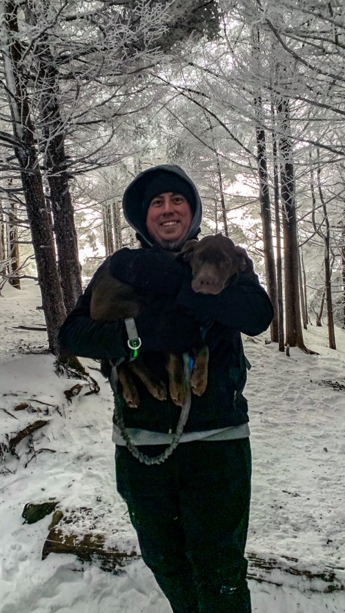 A person in winter attire holds a dog while standing on a snowy trail surrounded by frosty trees.