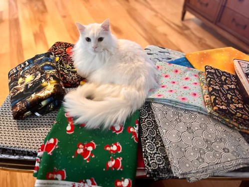 A fluffy white cat sits on a table surrounded by colorful fabric pieces in various patterns.