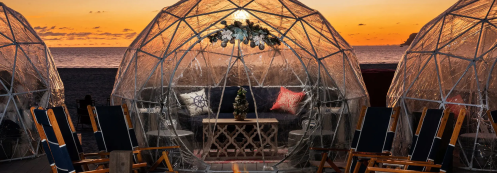 Three transparent igloos on a beach at sunset, featuring cozy seating and decorative elements.