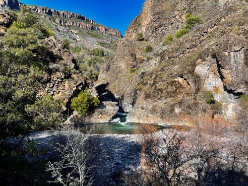 A serene river flows through a rocky canyon, surrounded by lush greenery and steep cliffs under a clear blue sky.