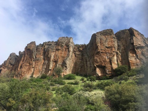 A rugged rock formation rises against a blue sky, surrounded by green vegetation and scattered clouds.