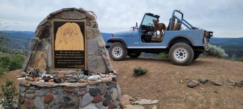 A blue Jeep parked on a rocky overlook with a dog inside, next to a stone monument and scenic mountain views.