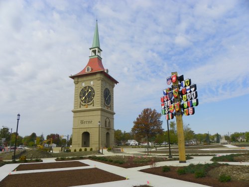 A clock tower with a green spire and a colorful shield display in a park setting under a cloudy sky.