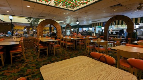 A spacious restaurant interior with wooden tables, orange chairs, and decorative arches, featuring a vibrant ceiling design.